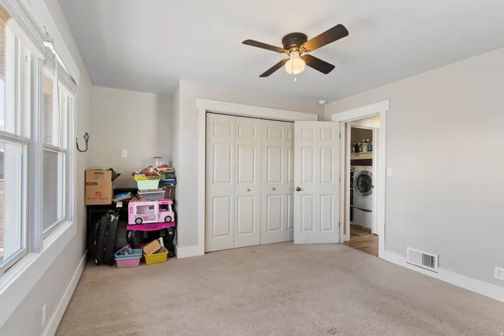Carpeted room featuring a ceiling fan, dual windows, bi-fold closet doors, and access to an in-unit laundry area with a visible washer