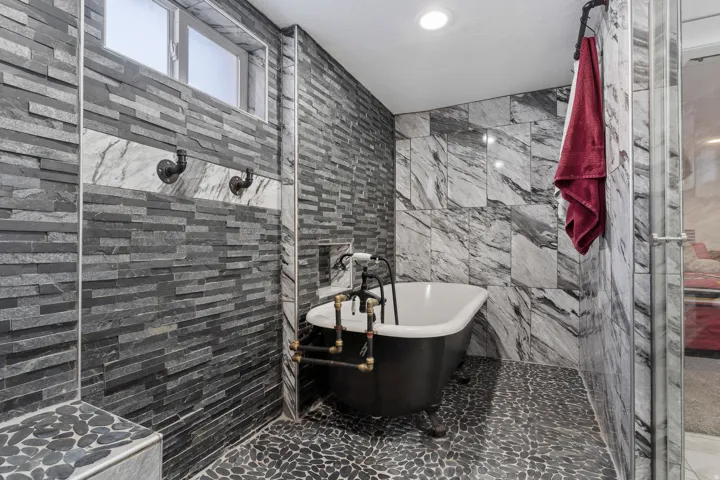 Freestanding clawfoot tub with exposed pipe-style fixtures, set against a backdrop of varied stone and tile finishes