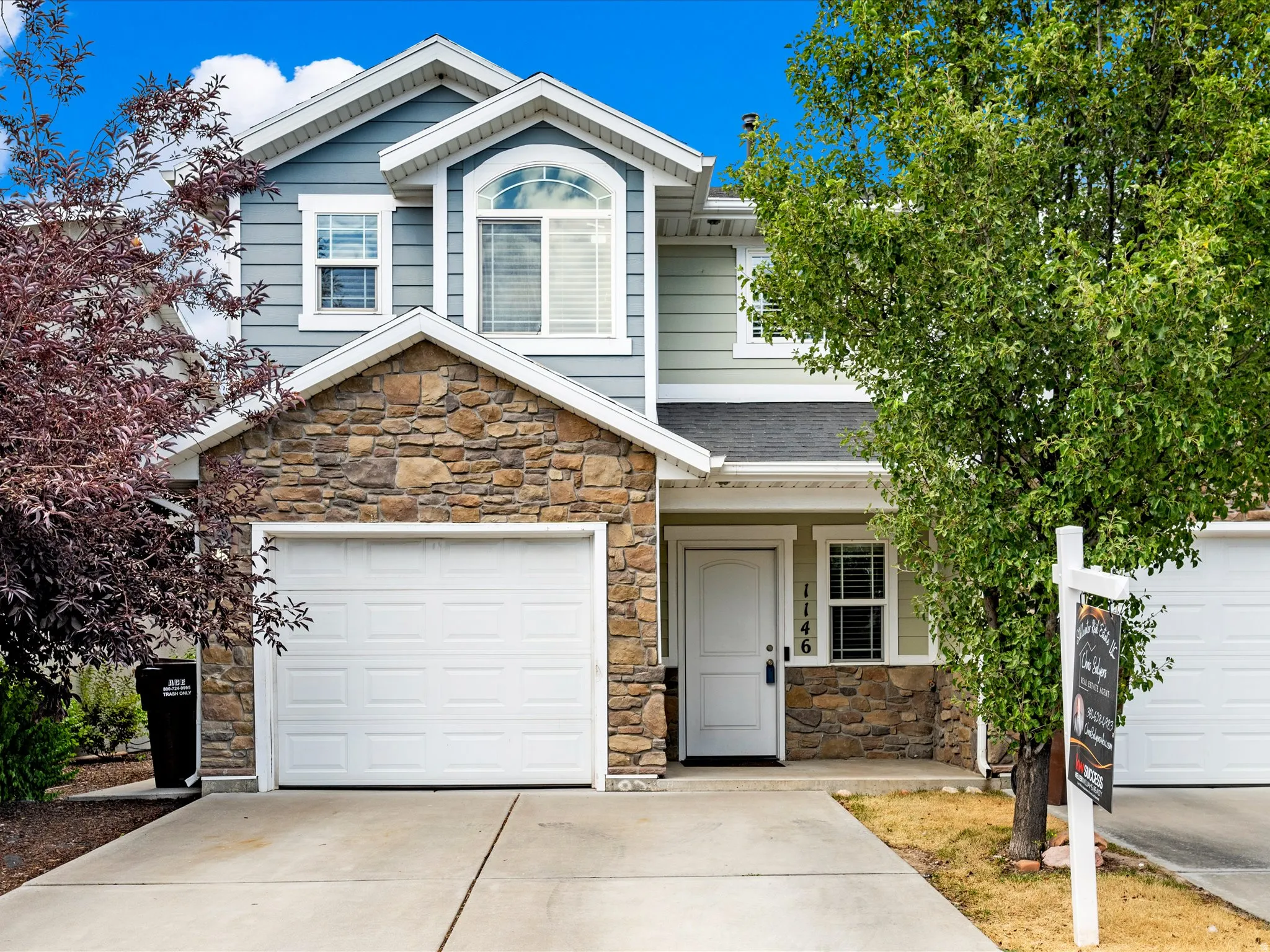 Two-story residence featuring a stone and siding facade, an arched window, an attached garage, a paved driveway, and mature landscaping
