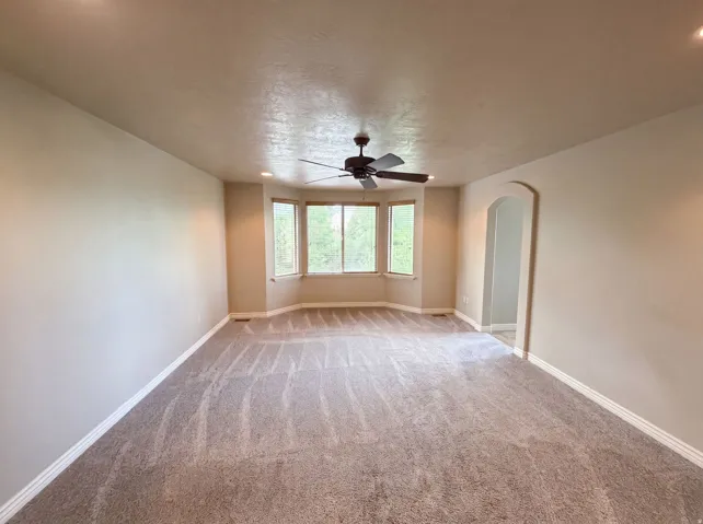 Spacious room featuring neutral carpeting, a multi-pane bay window, and a ceiling fan with integrated lighting