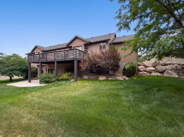 Rear exterior with stucco siding, a multi-level deck, a lower patio, mature landscaping, and a rock retaining wall