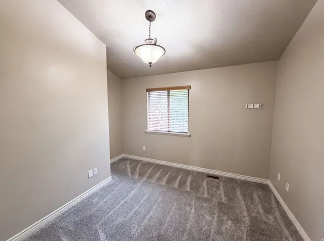 Neutral-toned walls complement the gray patterned carpet in this room featuring a ceiling-mounted light fixture, a window with horizontal blinds, and white baseboards