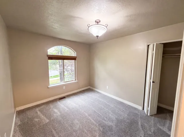 Room featuring an arched window with wood-finish blinds, textured ceiling, a flush-mount light fixture, gray patterned carpeting, and bi-fold closet doors