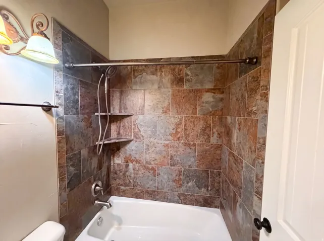 Bathtub with natural-toned tile surround, integrated shower, and corner shelving