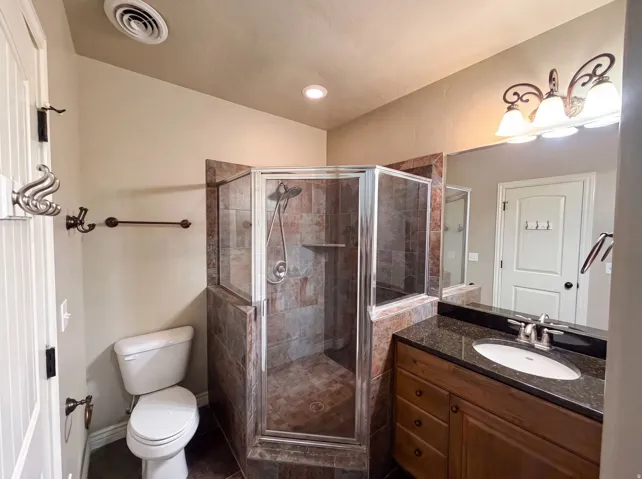 Bathroom featuring a corner glass-enclosed shower with tile surround, a single vanity with a dark countertop and undermount sink, a wall-mounted light fixture, and tile flooring