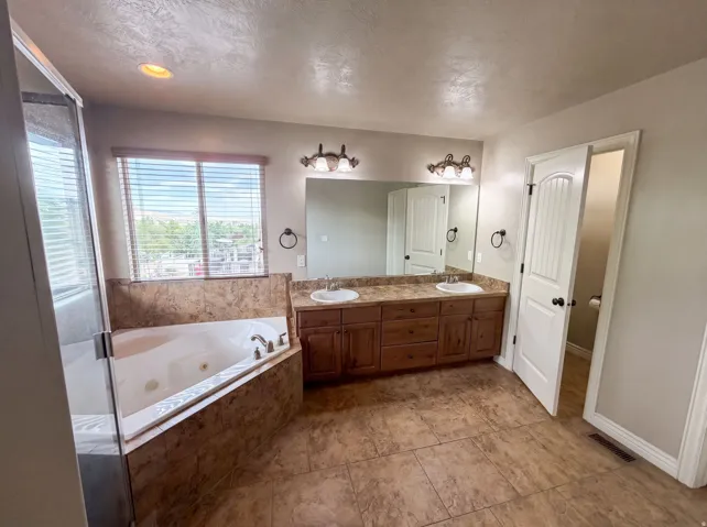 Bathroom featuring a corner jetted tub with tile surround, a spacious dual vanity with wood cabinetry and undermount sinks, and large-format floor tiling
