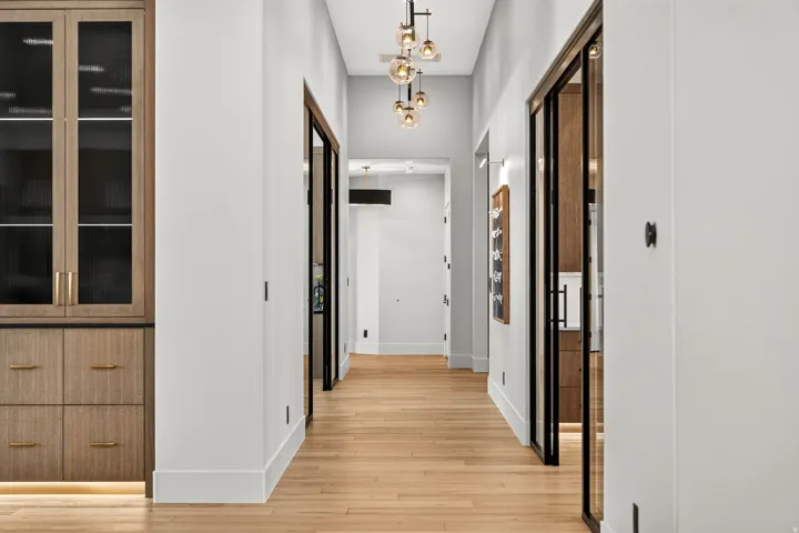 Hallway featuring wood-finish flooring, a contemporary multi-light pendant fixture, white walls, and dark-framed glass doors