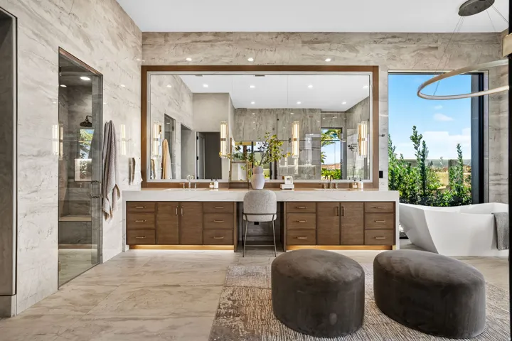 Spacious bathroom featuring a large frameless mirror, dual wood-finish vanity with stone countertop, freestanding soaking tub, and a glass-enclosed shower with a rainfall showerhead