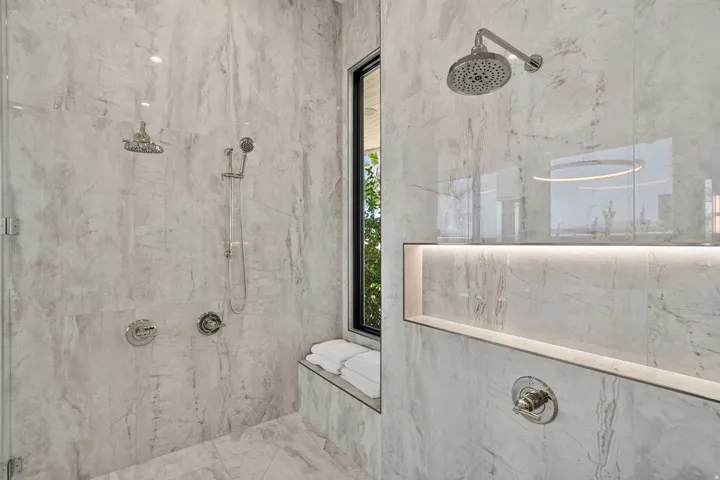 Spacious shower featuring light-toned stone-look tile, dual showerheads including a handheld sprayer, a built-in bench, a recessed niche with integrated lighting, and a black-framed window