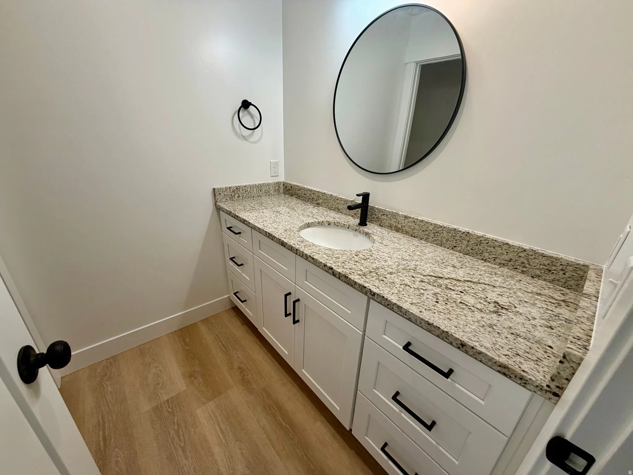 Bathroom vanity with white cabinetry, a light speckled countertop, and a circular black-framed mirror