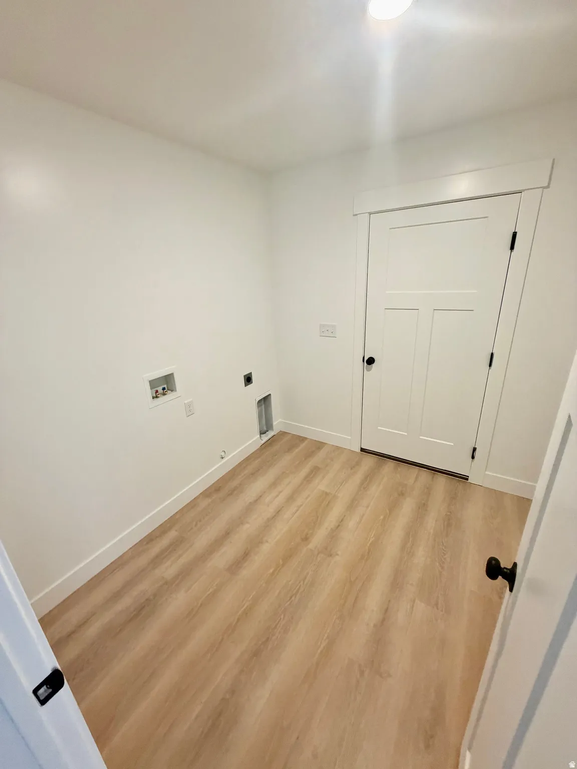 Laundry area featuring wood-finish flooring, white baseboards, a flush-mount ceiling light, and a panel-style door with black hardware
