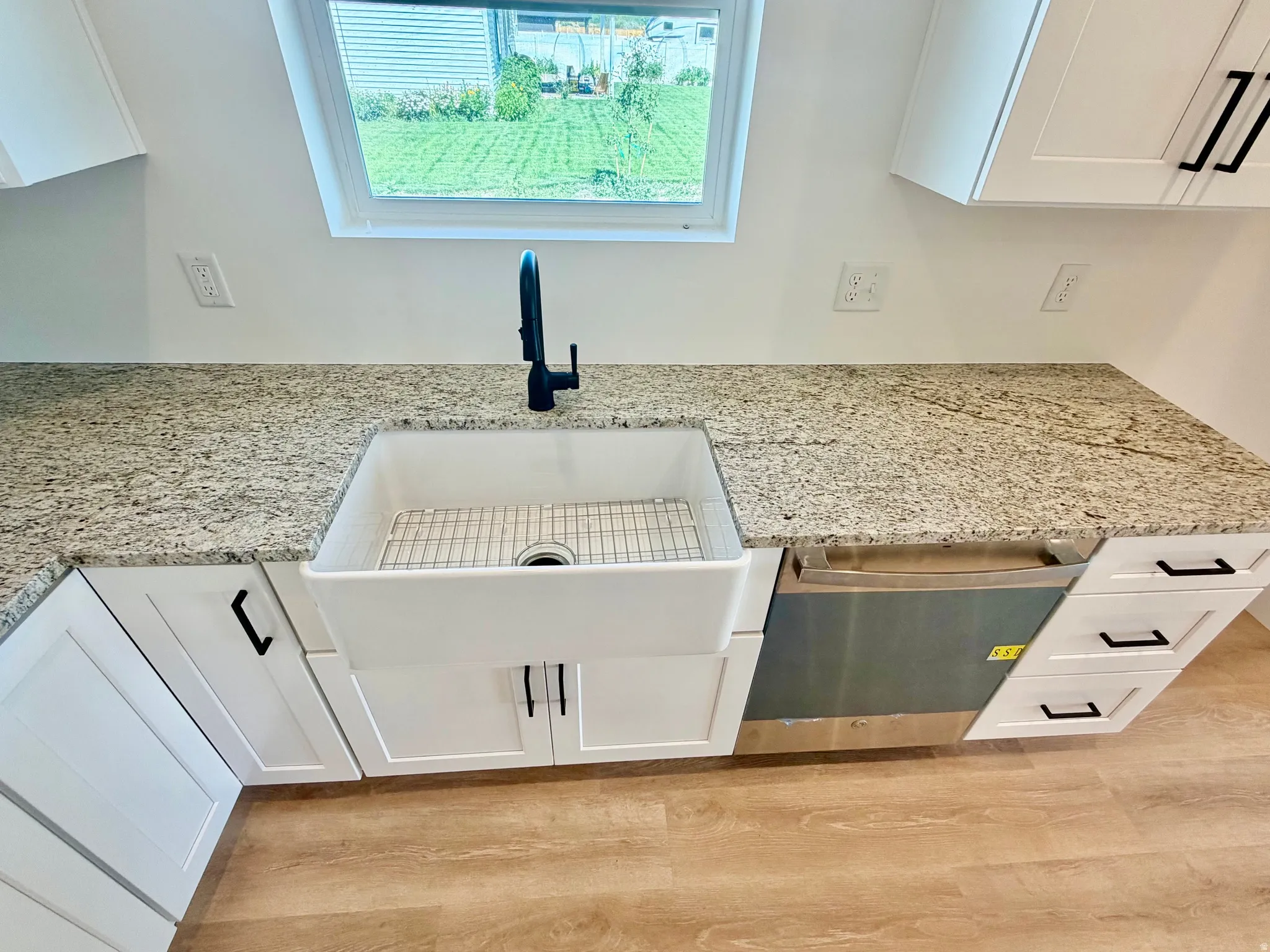 Kitchen featuring a white farmhouse sink with a matte black faucet, granite countertops, white shaker cabinetry with black hardware, and wood-finish flooring
