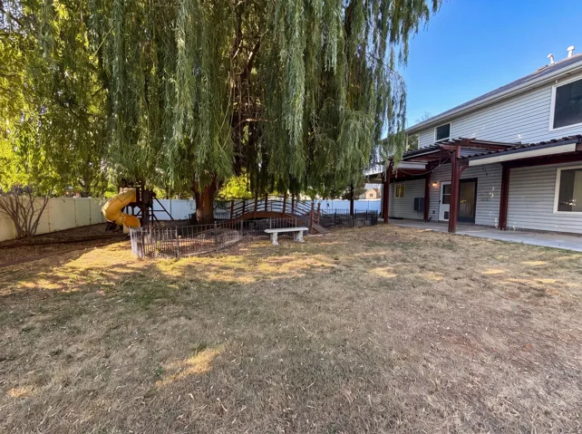 Expansive backyard featuring a large shade tree, a wooden pergola extending along the rear of the home, a white privacy fence, and a decorative bridge feature
