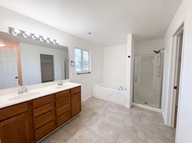 Bathroom featuring a dual vanity with wood cabinetry and white countertops, a large mirror with strip lighting, a built-in soaking tub, a glass-enclosed shower, and tile flooring