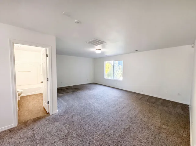 Spacious carpeted room featuring a ceiling-mounted light fixture, two windows, and an open doorway to a bathroom with tile flooring and a tub-shower combination