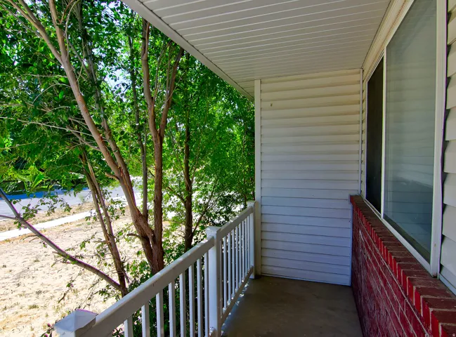 Covered porch with white vinyl siding, white railing, concrete flooring, and a red brick half-wall