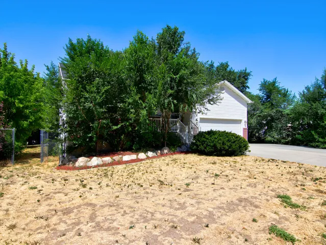 White siding exterior with an attached garage, a tiered front yard with natural rock landscaping, mature trees, and a chain-link fence