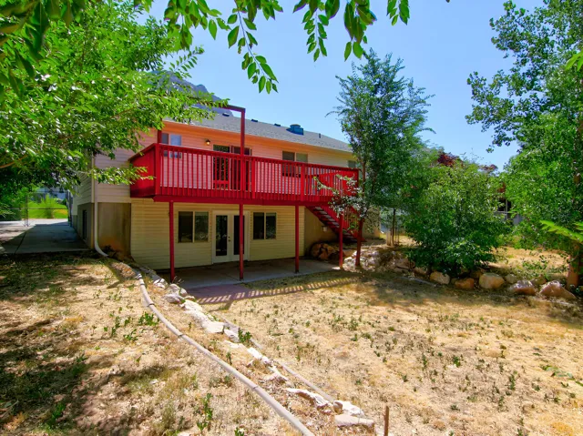 Rear exterior with a vibrant red elevated deck, multiple mature trees providing natural shade, a lower-level patio, and a tiered landscape