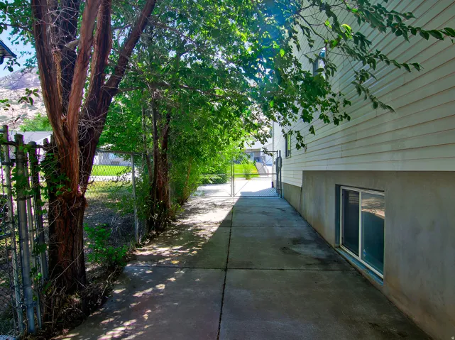 Concrete pathway alongside the property, featuring a chain-link fence, mature trees, and a sliding window on the lower level