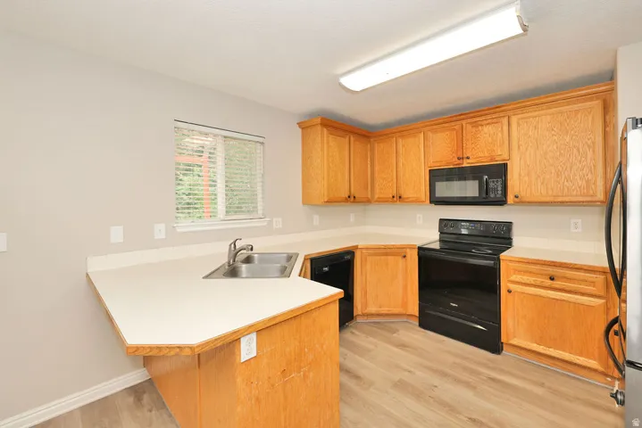 Kitchen featuring wood cabinetry, light countertops, black appliances, a double basin stainless steel sink, and wood-finish flooring