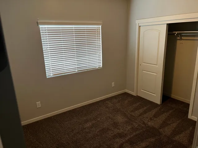 Neutral-toned room featuring a single window with blinds, brown patterned carpet, white baseboards, and a sliding closet door with an arched panel design