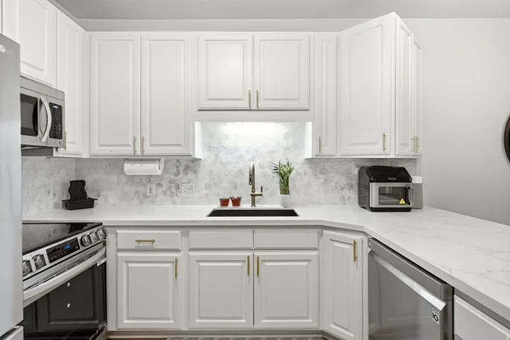 Modern kitchen featuring white shaker cabinetry with gold-tone hardware, stainless steel appliances, backsplash and quartz countertops.