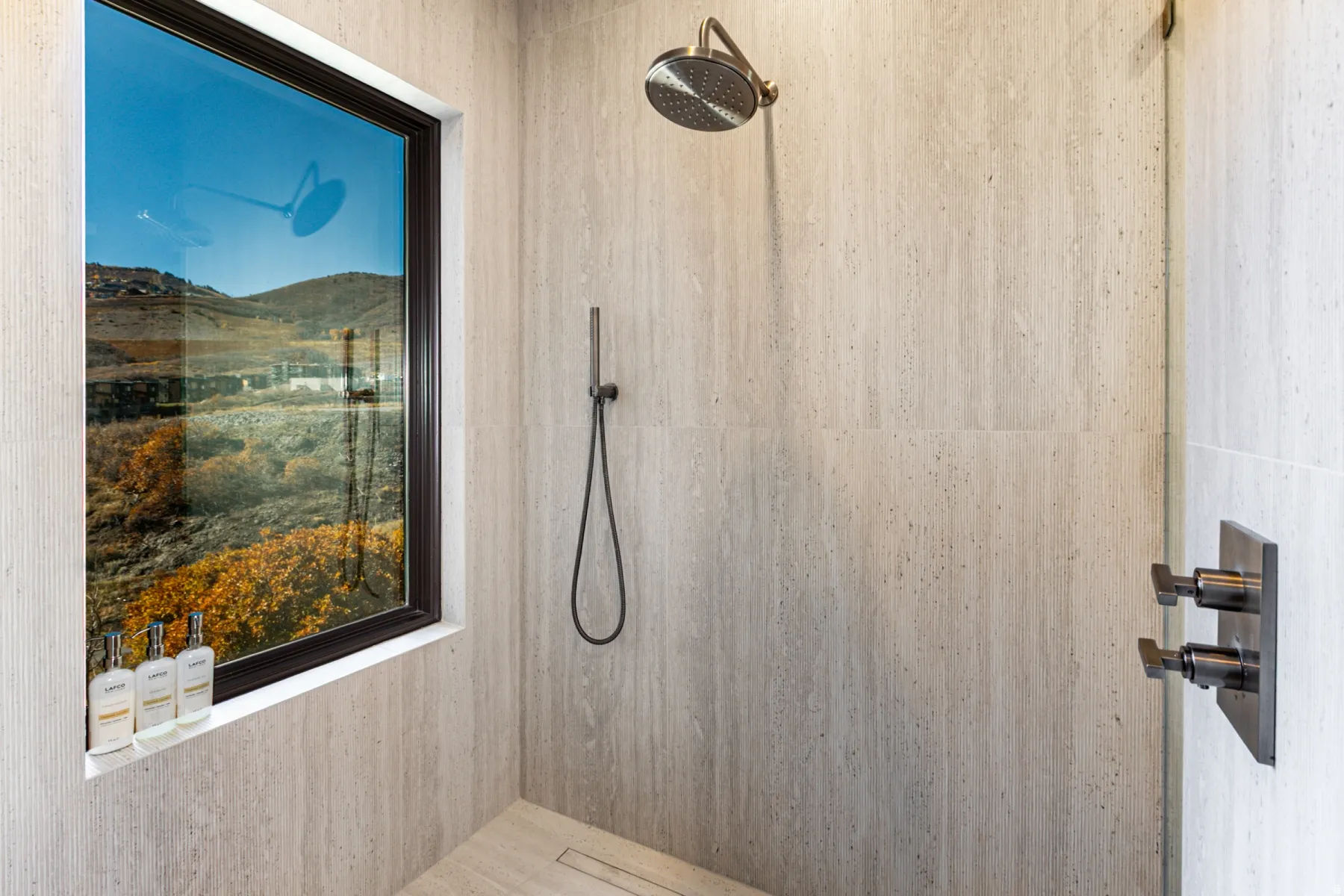 Shower with full-height window, light-toned stone-finish wall tile, bronze-finish fixtures, and a linear floor drain