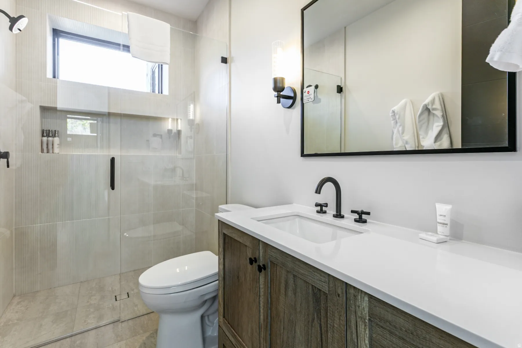 Contemporary bathroom featuring a glass-enclosed shower with a built-in niche, a wood-finish vanity with a white countertop, a rectangular undermount sink, and matte black fixtures