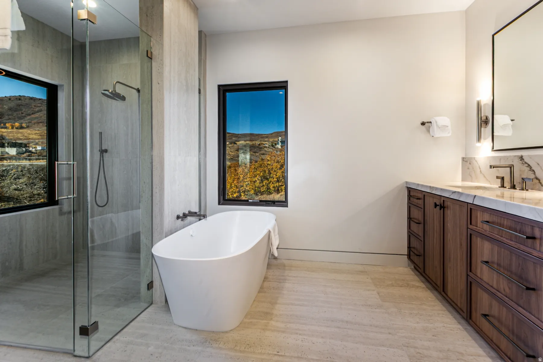 Modern bathroom featuring a freestanding soaking tub, a spacious glass-enclosed shower, a wood-finish vanity with a stone countertop, and two large windows