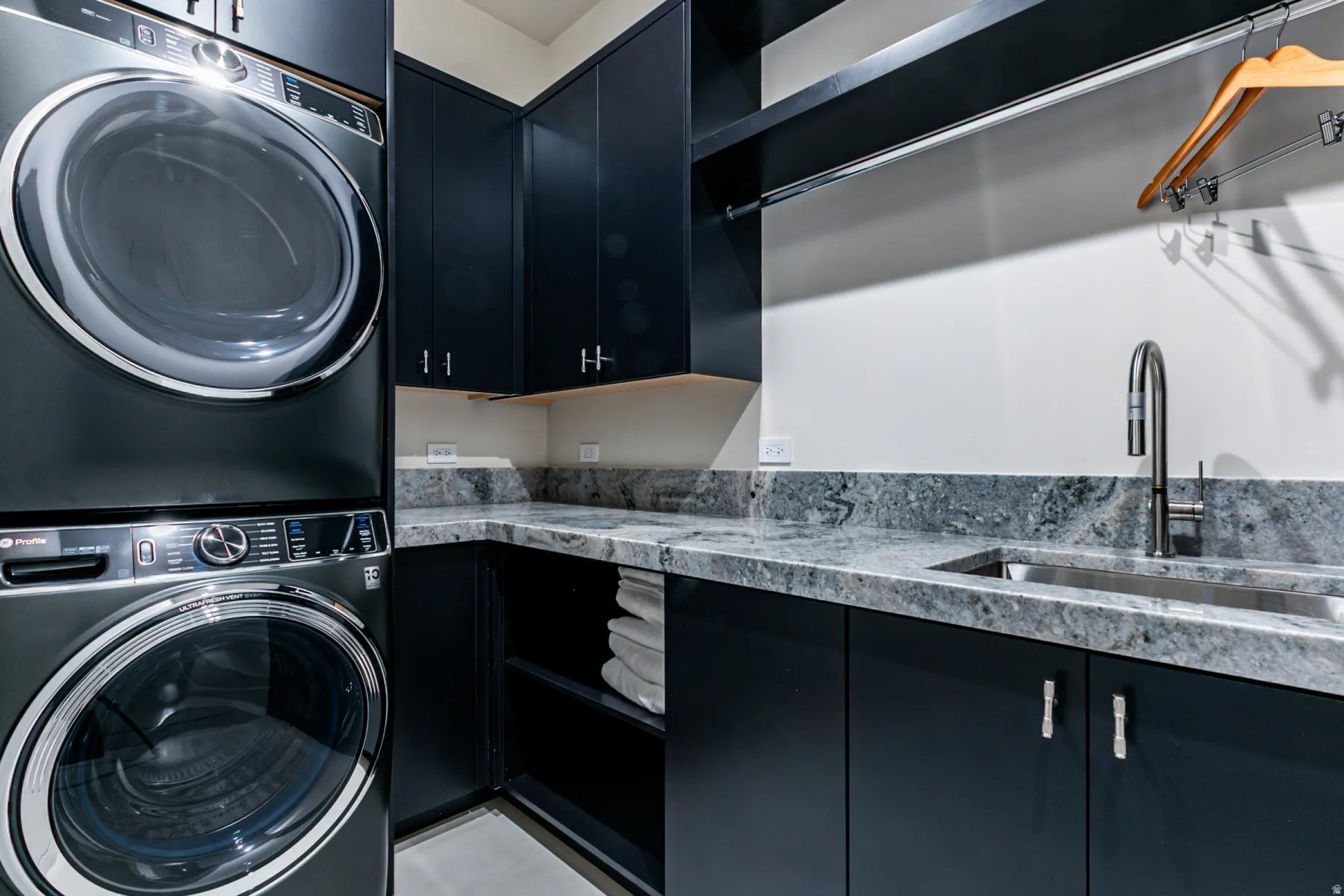 Laundry area featuring stacked washer and dryer, dark cabinetry with light-colored countertops, stainless steel sink with gooseneck faucet, and a hanging rod