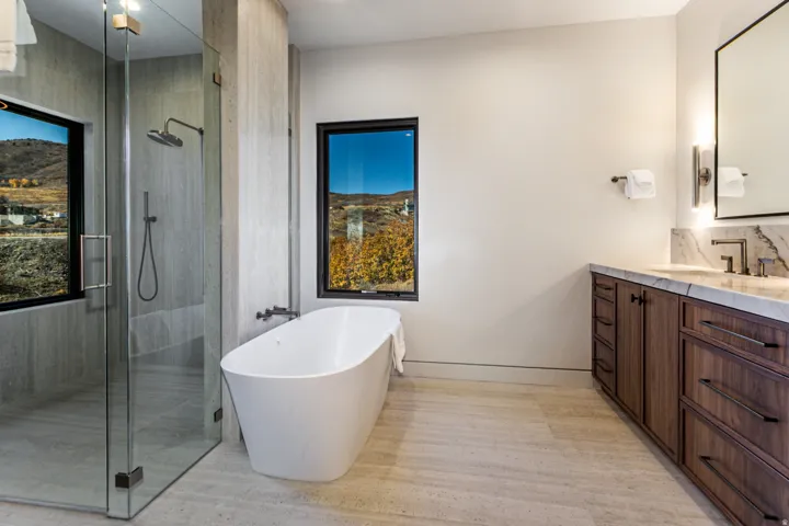 Modern bathroom featuring a freestanding soaking tub, a spacious glass-enclosed shower, a wood-finish vanity with a stone countertop, and two large windows