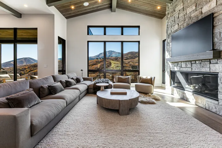 Vaulted wood-finish ceiling with exposed beams, stone fireplace, large floor-to-ceiling windows, and wood-finish flooring