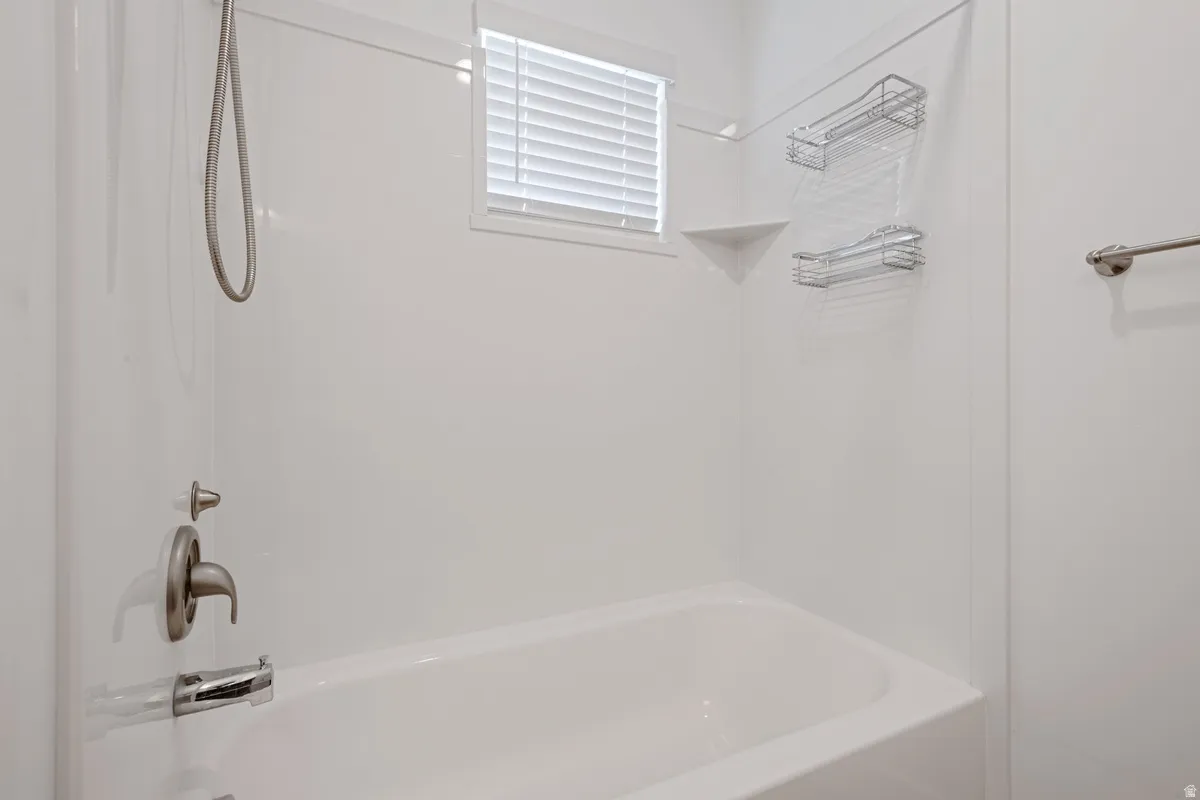 White bathtub and shower combination featuring brushed nickel fixtures, a window with horizontal blinds, and a built-in corner shelf