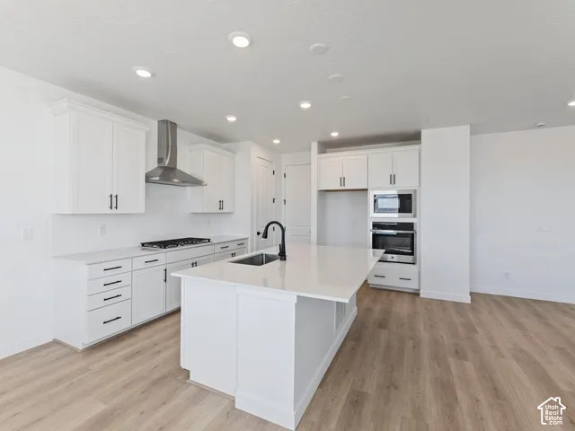 Modern kitchen featuring white cabinetry with hardware accents, a large waterfall-edge island with seating space, stainless steel appliances including a gas cooktop and wall-mounted oven, quartz countertops, and wood-finish flooring throughout the open la