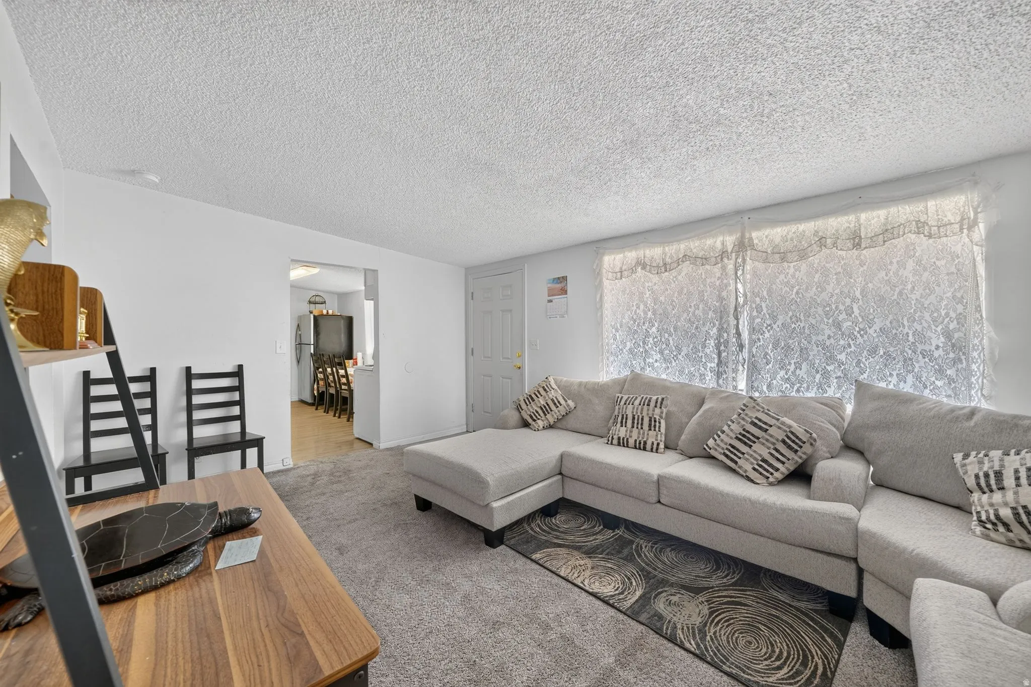 Carpeted living area featuring white walls, a textured ceiling, and a large window with lace coverings