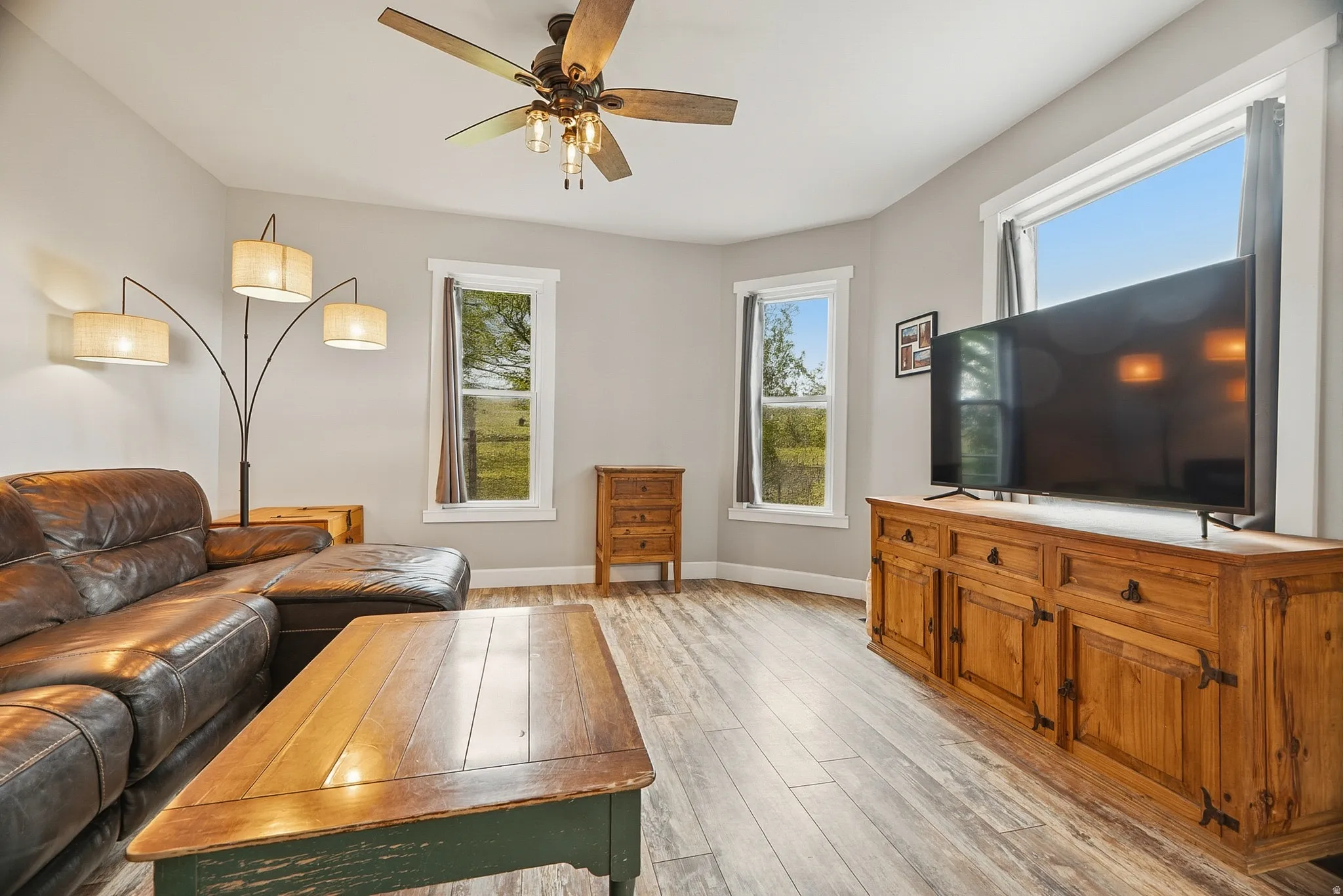 Living area featuring wood-finish flooring, a ceiling fan with integrated lighting, and multiple windows with white trim