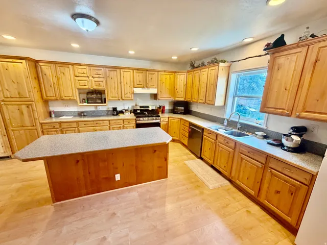 Spacious kitchen featuring extensive light wood cabinetry, light wood-finish flooring, a substantial center island with a light speckled countertop, recessed lighting, and a stainless steel double basin sink