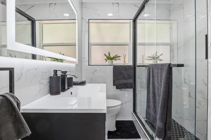 Contemporary bathroom featuring a black vanity with an integrated white sink, an illuminated framed mirror, and a glass-enclosed shower with black hexagonal tile flooring