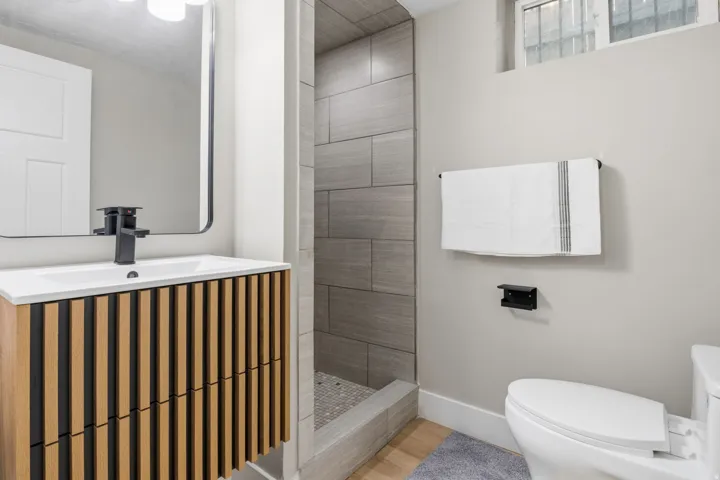Basement  Second Modern bathroom featuring a wood-panel vanity with a white countertop and rectangular sink, matte black faucet, and a frameless mirror
