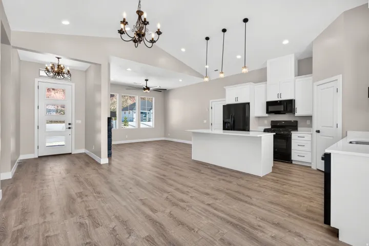 Open-concept living space featuring vaulted ceilings, wood-finish flooring, a kitchen island with pendant lighting, white cabinetry, and black appliances