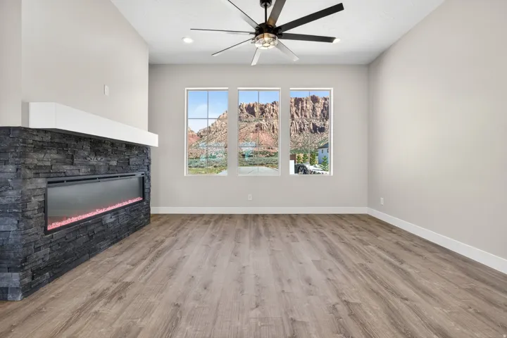 Spacious room featuring three large windows, a stone-clad linear fireplace with a white mantel, wood-finish flooring, and a modern ceiling fan