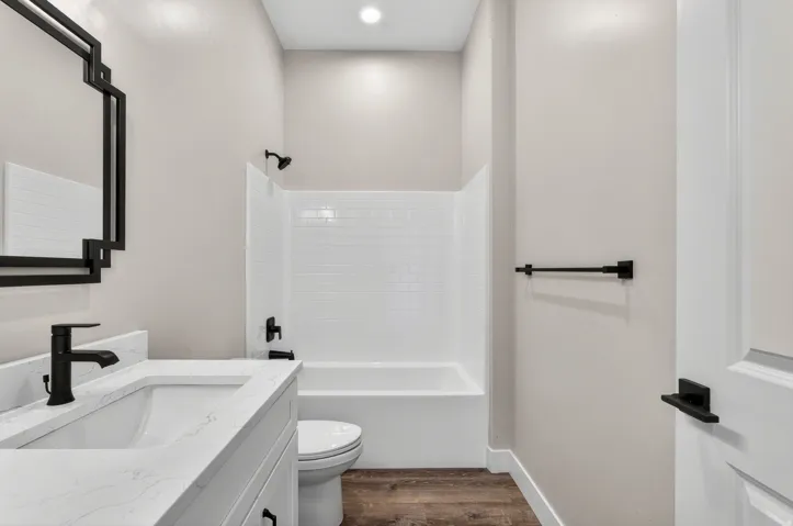 Bathroom featuring wood-finish flooring, a white vanity with an undermount sink and stone-finish countertop, a black framed mirror, a white bathtub with subway tile surround, and matte black fixtures
