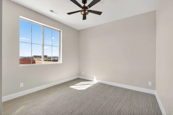 Room featuring a large window with gridded panes, a ceiling fan with dark blades and a bronze-toned fixture, light neutral-toned walls, and patterned carpeting
