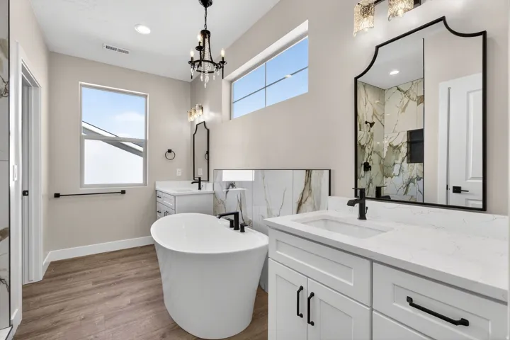 Spacious bathroom featuring a freestanding soaking tub, dual vanities with white countertops, wood-finish flooring, a contemporary black chandelier, and a walk-in shower with marble-look tile surround