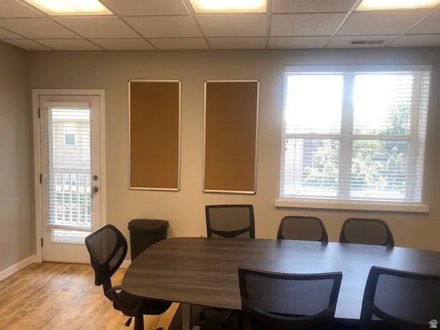 Room featuring wood-finish flooring, drop ceiling with integrated lighting, a white door with a glass panel and blinds, and a window with white blinds