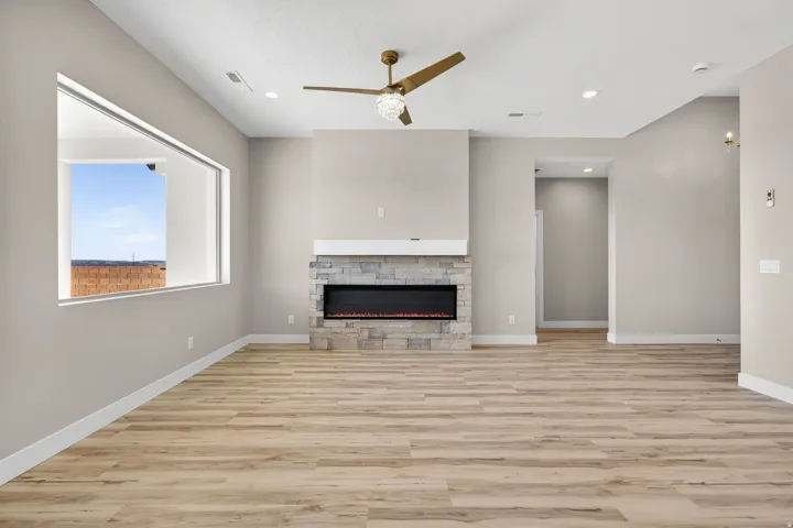 Spacious interior featuring wood-finish flooring, a contemporary fireplace with a stone surround and white mantel, a large horizontal window, a ceiling fan with integrated lighting, and recessed ceiling lights
