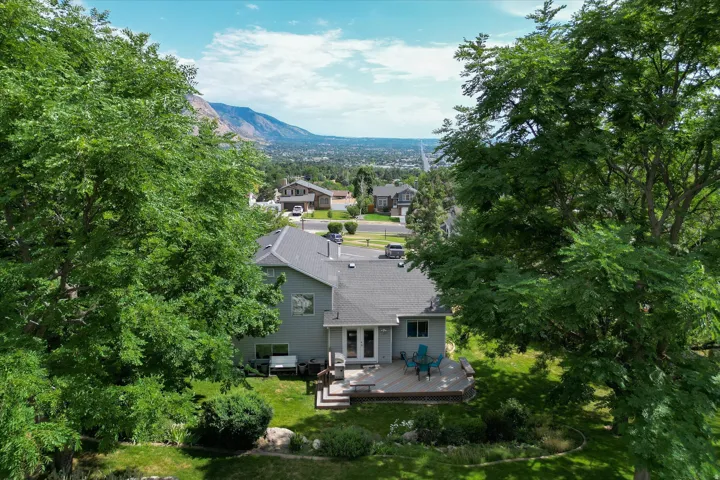 Rear exterior featuring a spacious deck with double doors, mature trees, manicured lawn, and distant mountains