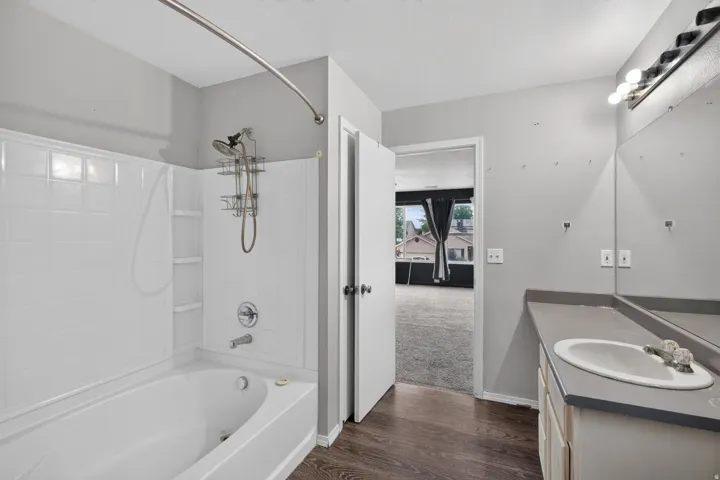 Bathroom featuring a built-in tub with a white tile surround, a single vanity with an integrated sink, a large mirror, and wood-finish flooring