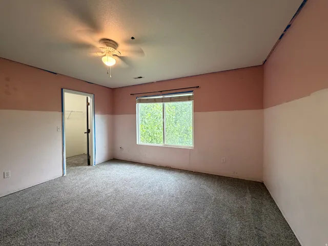 Carpeted interior room featuring a window, ceiling fan with light fixture, and an open doorway revealing a closet with wire shelving