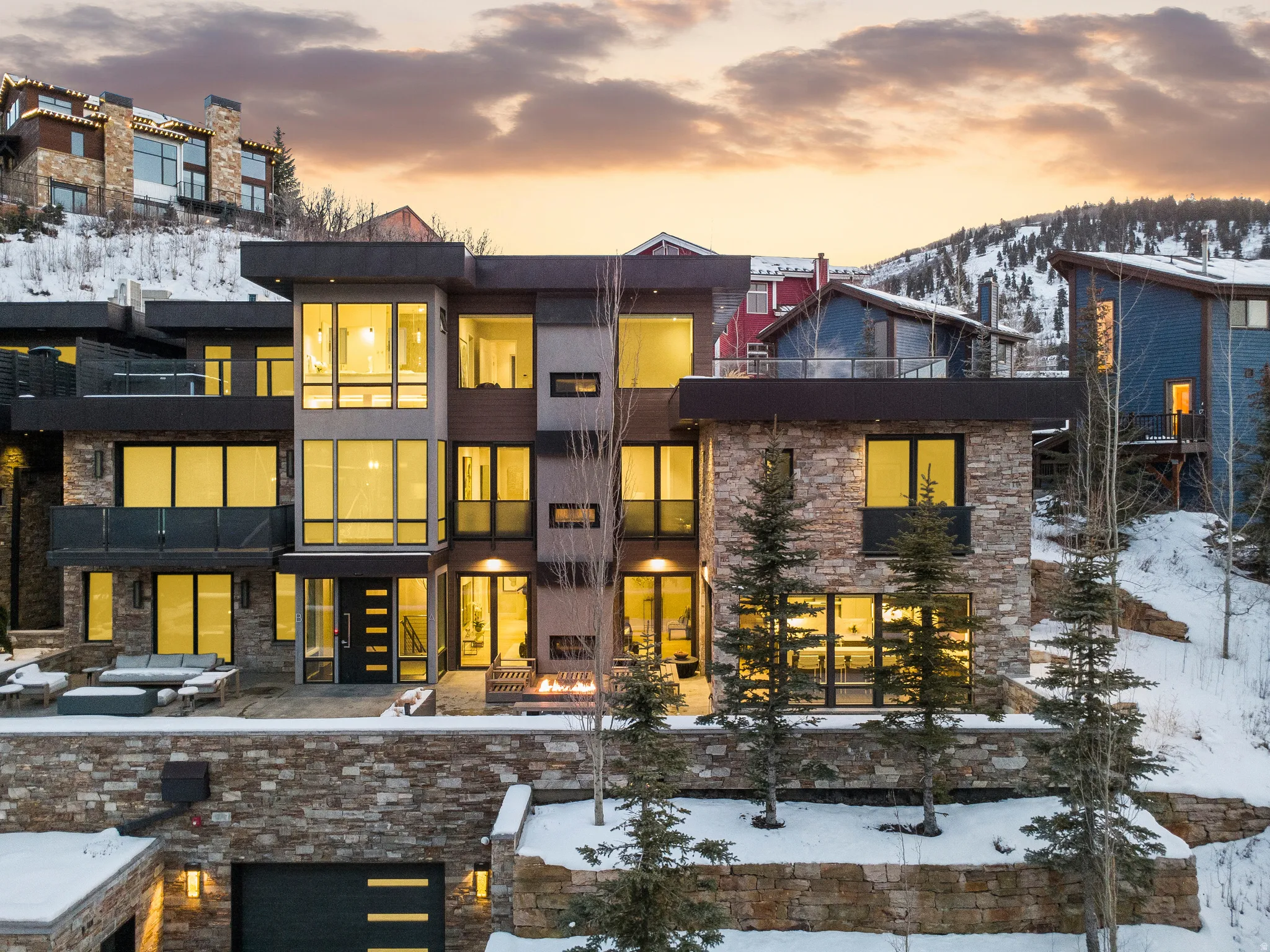 Contemporary multi-level residence featuring extensive stone and wood-finish siding, large format windows, a ground-level patio with a built-in fire pit, and an integrated garage